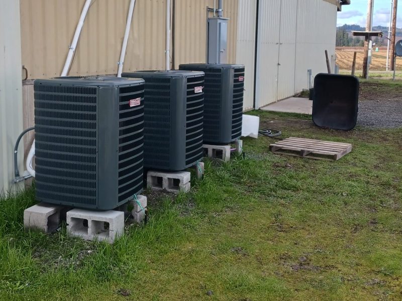 Three new AC condensing units installed on concrete block pads beside a metal barn