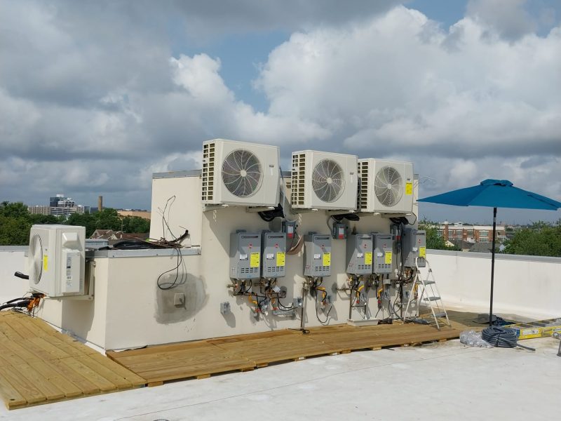 Rooftop installation of four heat-pump condensers above a bank of six tankless water heaters
